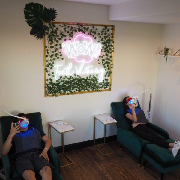 Two women sitting in recliners in a modern studio with a neon light behind them, both are receiving teeth whitening at the same time