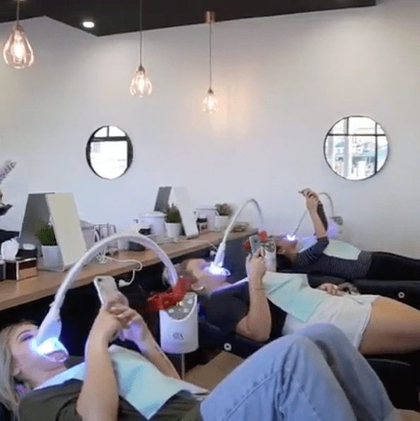 A group of 3 friends laying down receiving teeth whitening at the same time, all on their mobile devices.