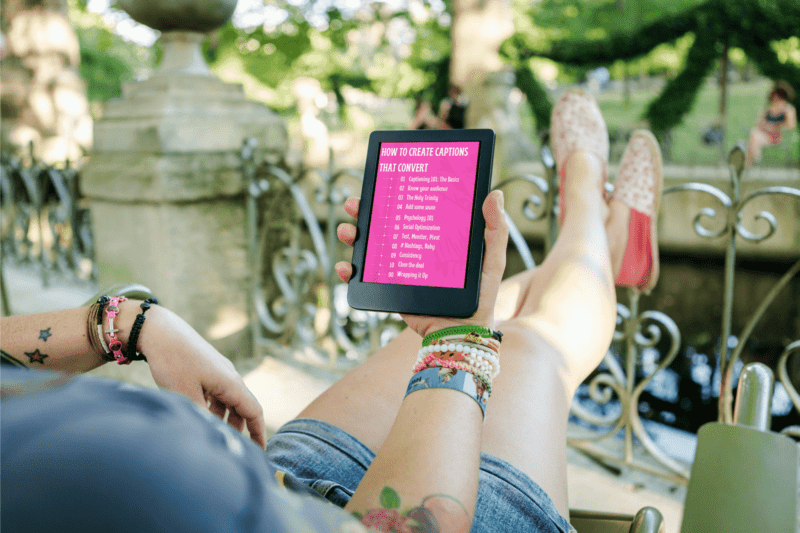 A woman sitting on her balcony with her feet reclined and an electronic device in her hand, reading the ebook. The woman appears hip and modern wearing bracelets and tattoos.